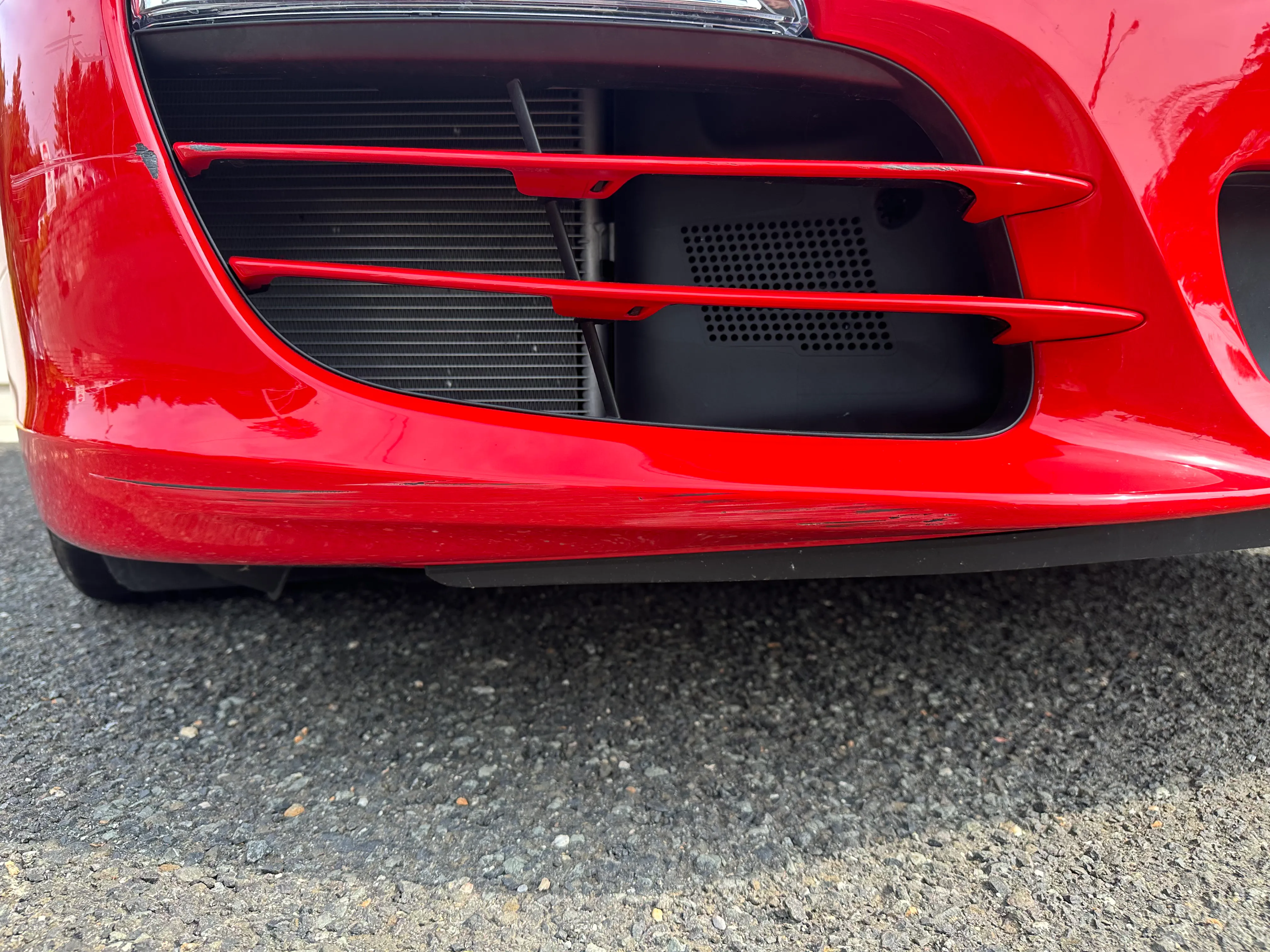 Red Porsche Boxster front bumper with scuff marks and scratches on the lower lip below the side intake, before repair.