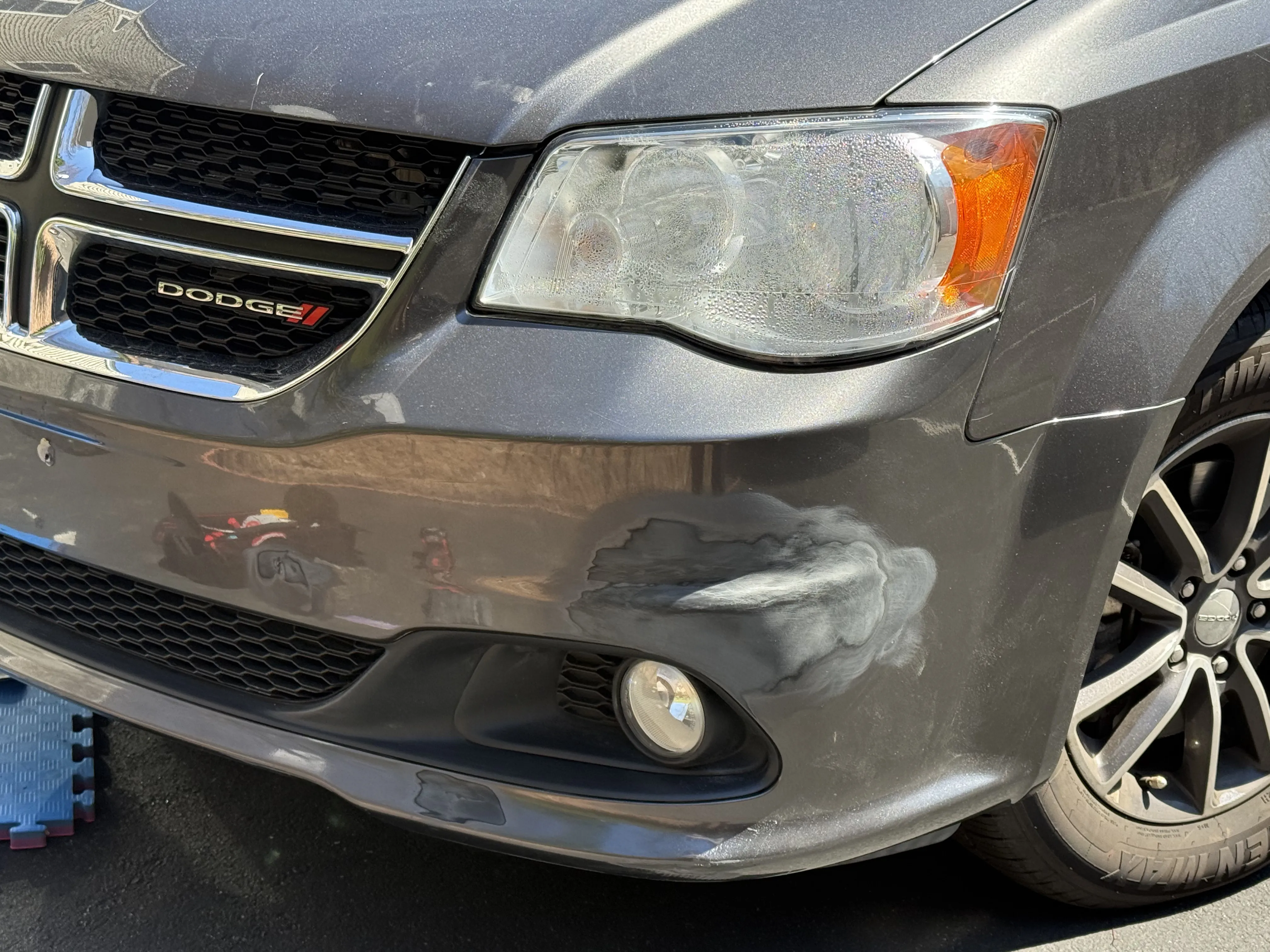 Gray Dodge Grand Caravan front bumper with visible horizontal scuffs and scratches below the driver-side headlight, before repair.