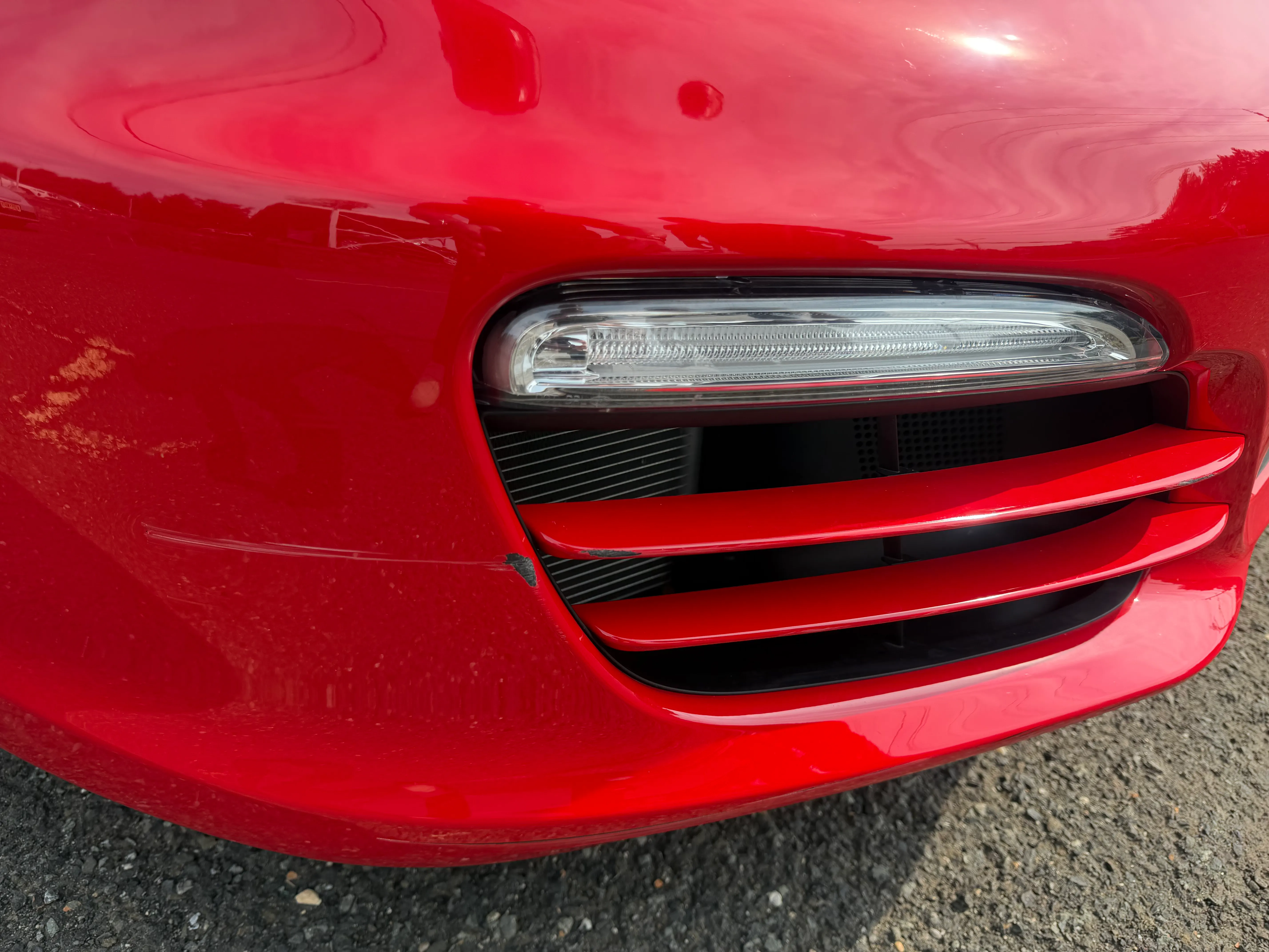 Close-up of the red Porsche Boxster front bumper side vent and running light, with a fine scratch and small chip visible beside the vent.